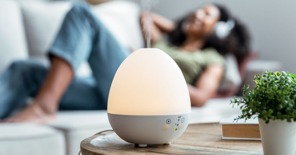 Shot of essential oil aroma diffuser humidifier diffusing water articles in the air while woman listening music lying on coach.