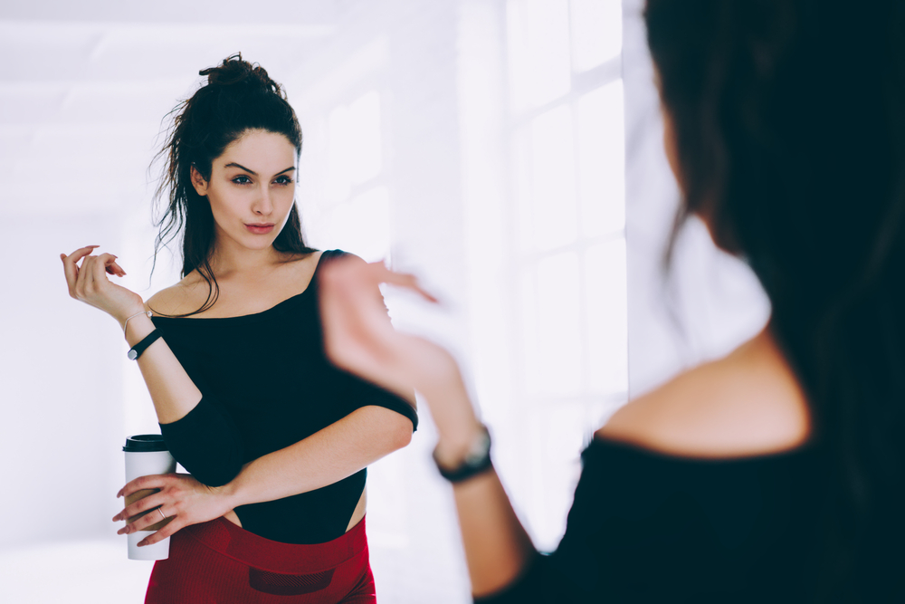 Brunette girl in tracksuit looking at mirror satisfied with body shape after workout in gym, narcissistic sportswoman checking muscles and figure in reflection care about healthy lifestyle